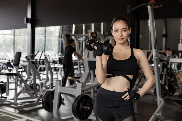 Ejercicio de mujeres sexy asiáticas con pesas en el gimnasio con espacio de copia. Linda mujer con mancuernas de elevación. Muchacha tailandesa que sostiene la pesa de gimnasia en el fondo del gimnasio. Concepto de ejercicio en el gimnasio.