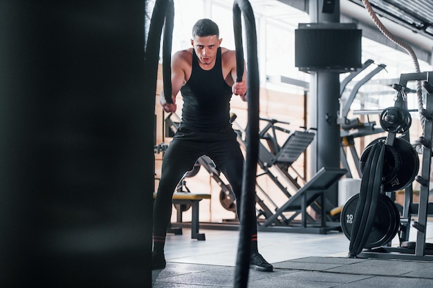 Ejercicio con nudos Joven deportista fuerte vestido de negro tiene un día de entrenamiento en el gimnasio