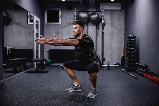 Ejercicio para quemar piernas con la nueva tecnología EMS Un hombre joven y atractivo vestido con ropa EMS haciendo sentadillas