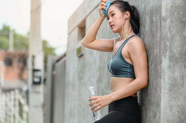 Ejercicios deportivos cansados de la mujer asiática. Ella usa una toalla limpia el sudor de su rostro. Ella está de pie al aire libre. Fitness, entrenamiento, ejercicio de gimnasio, estilo de vida y concepto saludable.