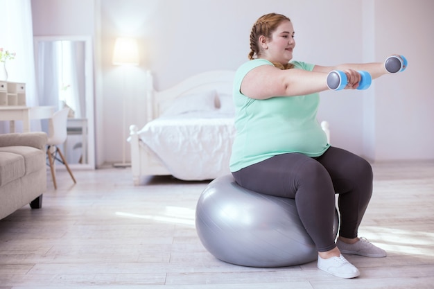 Ejercicios con pelota. Mujer sonriente regordeta sentada sobre la pelota mientras hace ejercicios con pesas