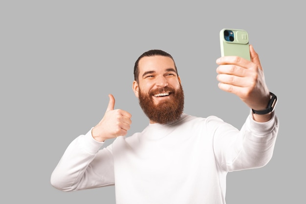 Ekstatisch erstaunter bärtiger Mann hält Telefon und macht ein Selfie damit, während er den Daumen nach oben zeigt