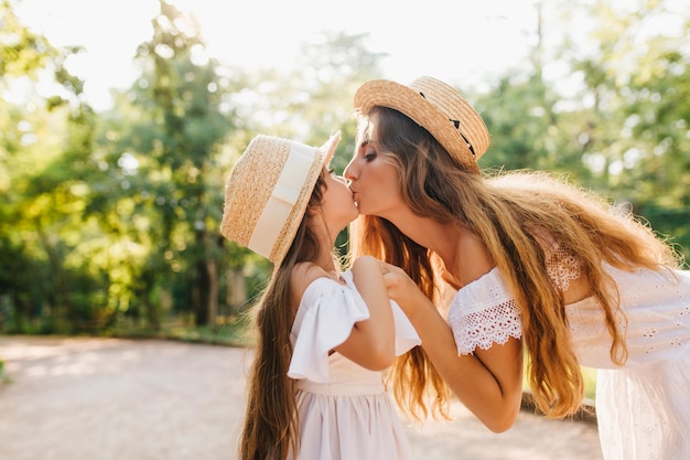 Ekstatische Frau mit langen glänzenden Haaren, die ihre Tochter während der Freizeit im Park sanft küsst. Wunderschöne junge Dame, die Händchen mit kleinem Mädchen hält und ihre Augen auf Naturhintergrund untersucht.