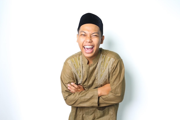 Ekstatischer asiatischer muslimischer Mann, der mit verschränkten Armen lacht und islamische Kleidung trägt, isoliert auf weißem Hintergrund