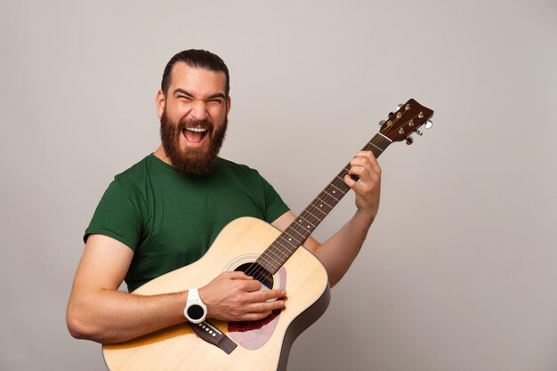 Ekstatischer junger bärtiger Mann hält und spielt Rock auf einer Akustikgitarre