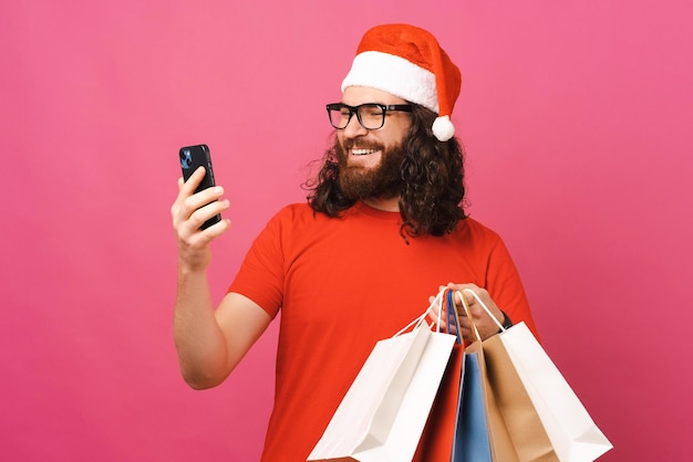 Ekstatischer Mann mit Weihnachtsmütze kauft am Telefon ein, während er einige Taschen hält