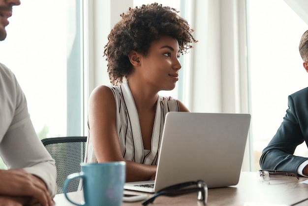 Ela adora seu trabalho. Linda mulher africana sentada na reunião de negócios e sorrindo. Equipe multicultural