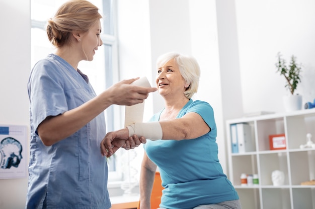 Elastische Bandage. Angenehme, erfreute Krankenschwester, die ihren Patienten ansieht, während sie einen weißen Verband hält holding