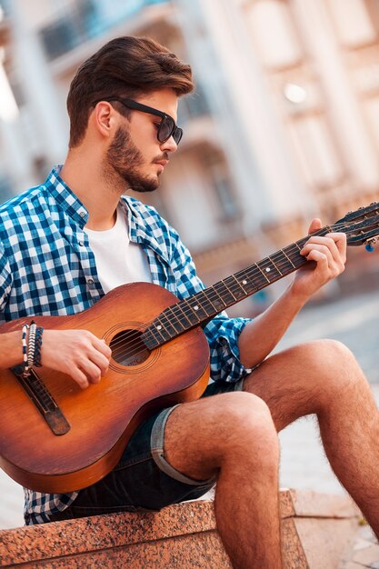 Ele adora esse som. Vista de baixo ângulo de um jovem bonito tocando violão enquanto sentado ao ar livre