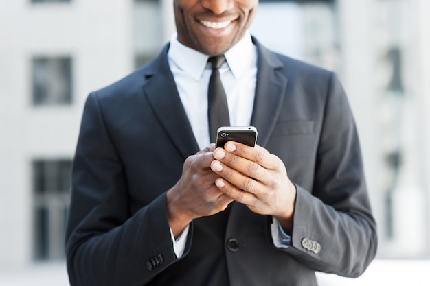 Ele está sempre em contato. Imagem recortada de um jovem africano alegre em trajes formais segurando um telefone celular e sorrindo ao ar livre