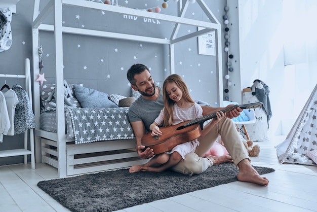 Ele sempre acredita nela. Jovem pai ensinando sua filha a tocar violão e sorrindo