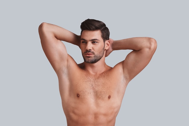 Ele tem um torso perfeito. Jovem bonito, mantendo as mãos atrás da cabeça e olhando para longe em um fundo cinza