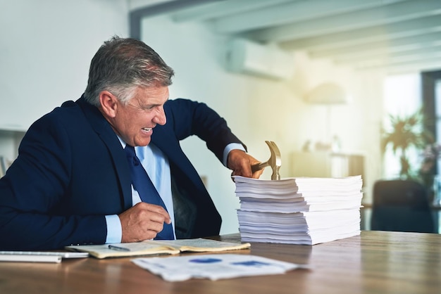 Ele tem uma abordagem alternativa para a papelada Foto de um empresário frustrado batendo em uma pilha de papelada com um martelo