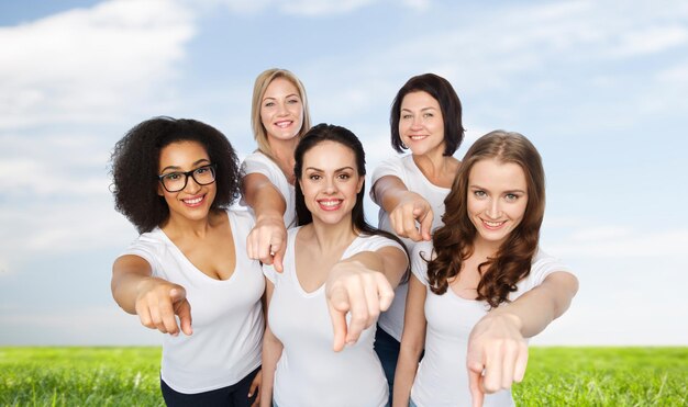 elección, amistad, cuerpo positivo, gesto y concepto de personas - grupo de mujeres felices de diferentes tamaños con camisetas blancas señalándote con el dedo sobre el cielo azul y el fondo de la hierba