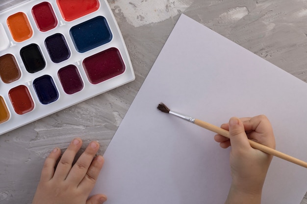 Elección de colores para la primera pintura: primer plano de cubos de pintura con pincel y mano para niños, vista superior