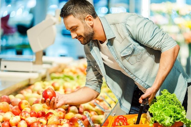 La elección nutricional. Apuesto joven sosteniendo manzana y bolsa de compras mientras está de pie en una tienda de alimentos