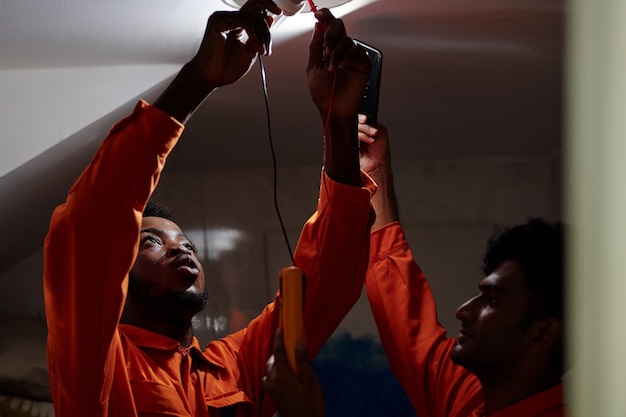 Electricista afroamericano en traje naranja midiendo la energía eléctrica mientras inserta la bombilla con un compañero de trabajo sosteniendo el teléfono con linterna