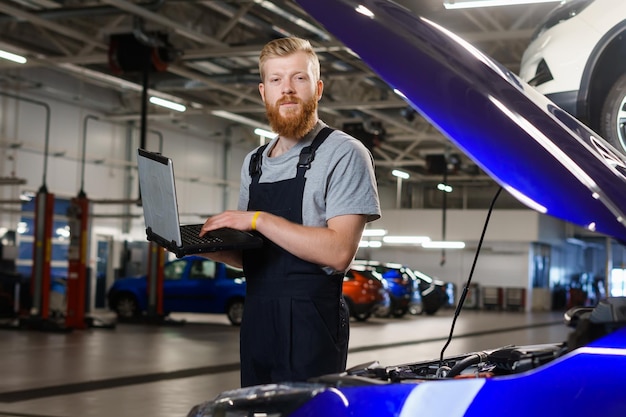 Un electricista automotriz masculino se para con una computadora portátil de diagnóstico en el automóvil mientras trabaja como empleado de la estación de servicio