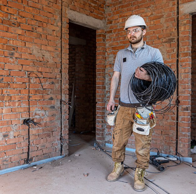 Un electricista con casco mira la pared mientras sostiene un cable eléctrico.