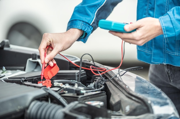 El electricista o mecánico de automóviles comprueba el voltaje en la batería del automóvil dentro del motor del automóvil con el capó abierto.
