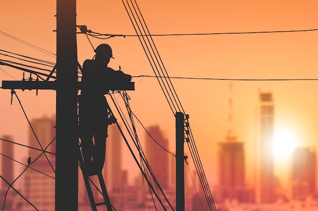 Electricista de silueta instalando líneas de cable en poste eléctrico con fondo de cielo de amanecer borroso