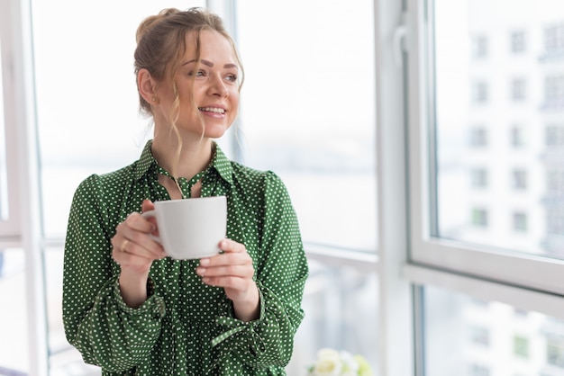 Elegancia hermosa mujer rubia en un rascacielos istanding sosteniendo una taza en el fondo de la ventana