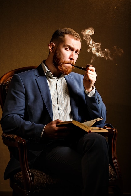 Elegancia y masculinidad. Retrato de joven guapo en traje azul fumando una pipa y leer el libro, sentado en la silla.