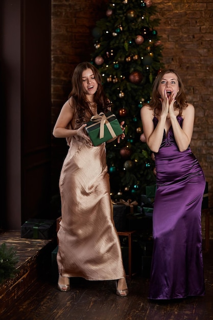 Elegancia mujeres delgadas en vestidos de noche dando una caja de regalo cerca del árbol de Navidad Concepto de día de boxeo de Nochevieja 39 Hermanas felices o novias divirtiéndose juntas Vacaciones buen humor