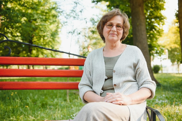 Elegante ältere Frau im Hemd sitzt an einem warmen Tag auf der Bank in einem Park