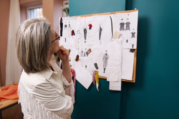 Elegante anciana europea mirando cuidadosamente un stand con bocetos de ropa anclados y esquises de pie en su propio taller de diseño de moda