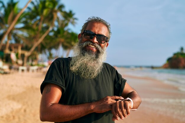 El elegante anciano de barba gris con gafas de sol usa un reloj inteligente en una playa tropical.