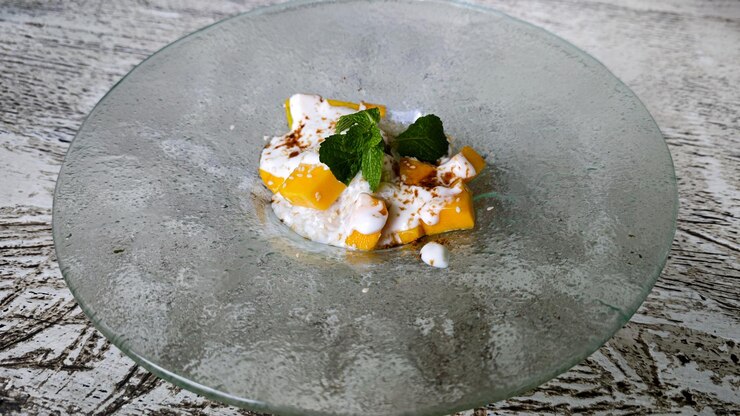 Foto elegante apresentação de arroz pegajoso de manga com creme de coco em um prato de vidro com folhas de hortelã enfeitadas em um ambiente rústico brilhante
