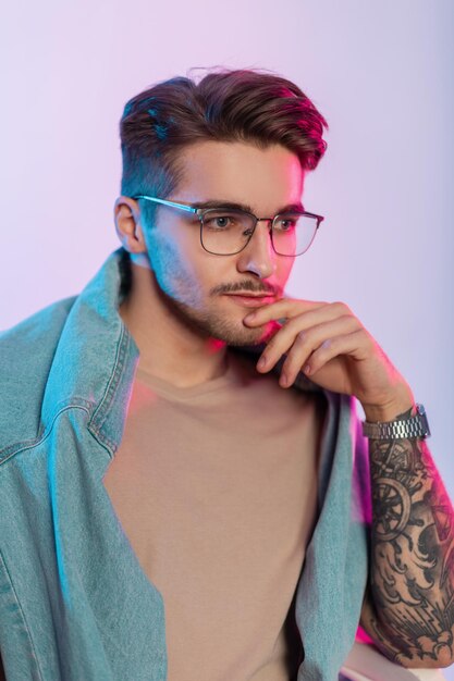 Elegante, apuesto y creativo artista con corte de pelo y anteojos antiguos con ropa informal de moda con tatuajes en el brazo y horas sentado en el estudio con luces rosas