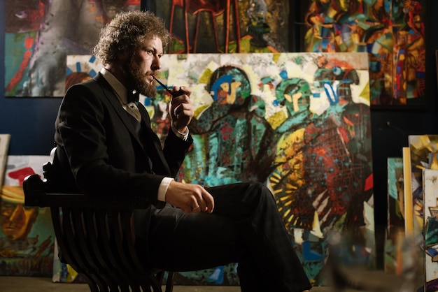 Elegante artista pintor barbudo fumando una pipa cerca de sus pinturas