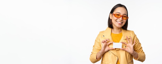 Elegante y atractiva chica asiática con gafas de sol mostrando tarjeta de crédito y sonriendo de pie feliz contra el fondo blanco del estudio