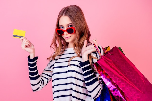 Elegante y atractiva mujer joven con un suéter a rayas y gafas de sol rojas tiene muchas bolsas de la compra y tarjetas de crédito sobre un fondo rosa en el estudio. concepto de adicción a las compras y venta.