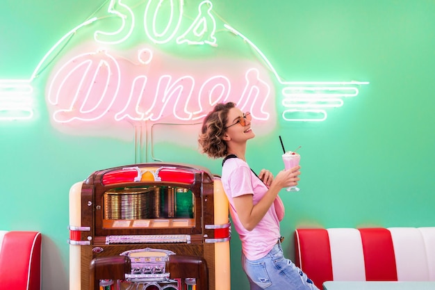 Elegante y atractiva mujer sonriente en un café retro de los años 50 en una jukebox de música bebiendo un cóctel de batido de leche con ropa hipster divirtiéndose riendo de buen humor