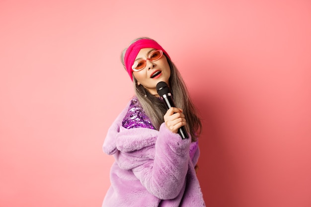 Elegante y atrevida mujer madura asiática realiza una canción en el escenario, sosteniendo el micrófono y cantando karaoke, con gafas de sol de moda en forma de corazón y un abrigo de piel sintética, fondo rosa