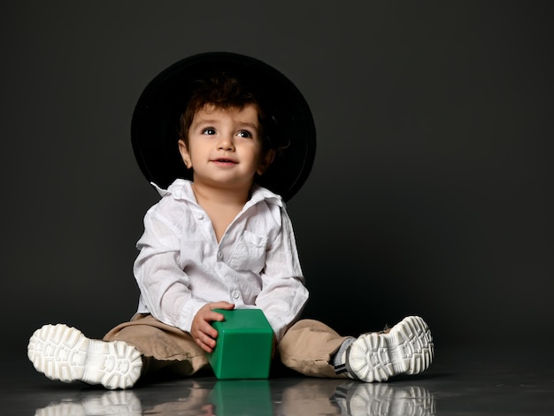 Elegante bebê menino criança modelo usando sapatos de vestuário casual e chapéu segurando o bloco de construção de brinquedo Retrato isolado isolado tiro no fundo cinza do estúdio Roupa na moda para propaganda infantil