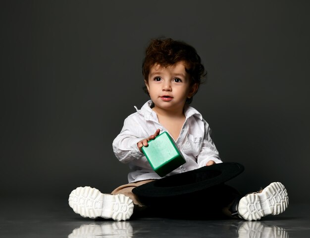 Elegante bebé niño Niño pequeño modelo vistiendo ropa casual zapatos poniendo bloque de construcción de juguete en sombrero Retrato aislado tiro aislado sobre fondo gris de estudio Traje de moda para anuncio de niño
