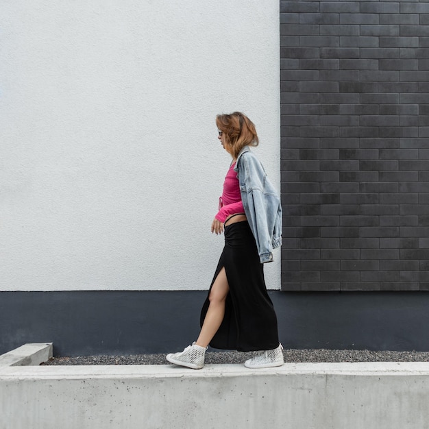 Elegante bela jovem ruiva modelo mulher com óculos de sol brancos em roupas de rua da moda com uma jaqueta jeans de saia longa de moletom rosa e tênis andando perto de uma parede de tijolo branco e preto