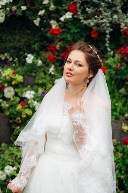 Elegante Braut steht auf dem Hintergrund eines Blumengartens in einem eleganten Hochzeitskleid und einem Brautschleier mit einem Diadem