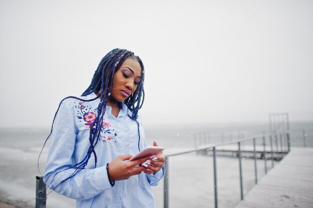 Elegante chica afroamericana con rastas sosteniendo el teléfono móvil a mano, al aire libre en el muelle contra el lago congelado en clima nevado.