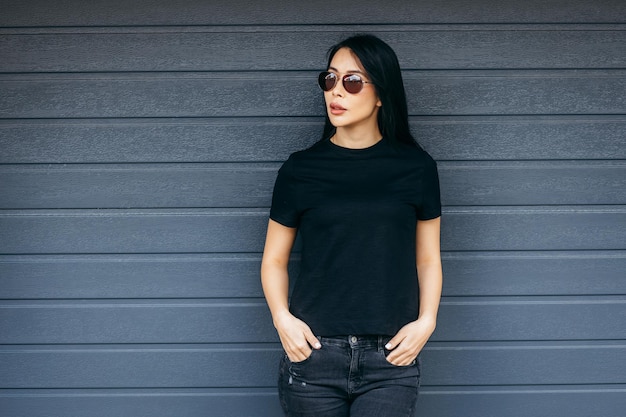 Elegante chica asiática morena con camiseta negra y gafas de sol posando contra la tela urbana de la calle