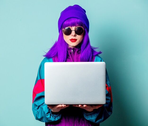 Elegante chica blanca con cabello morado y chándal con laptop en pared azul