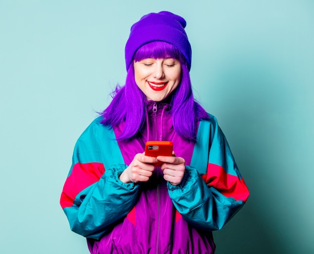 Elegante chica blanca con pelo morado y chándal mediante teléfono móvil en la pared azul