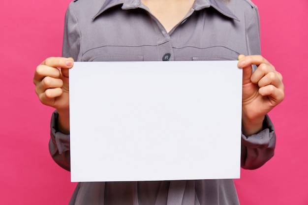 Foto elegante chica caucásica con un vestido gris sobre un fondo rosa. sostiene un trozo de papel blanco. copia espacio