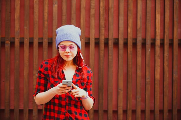 Elegante chica hipster informal escucha música usando un teléfono inteligente y auriculares sobre una pared de madera