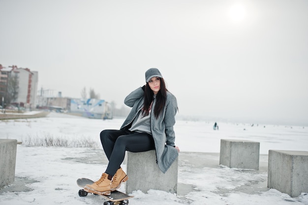 Elegante chica morena con gorra gris estilo casual street con patineta en el día de invierno