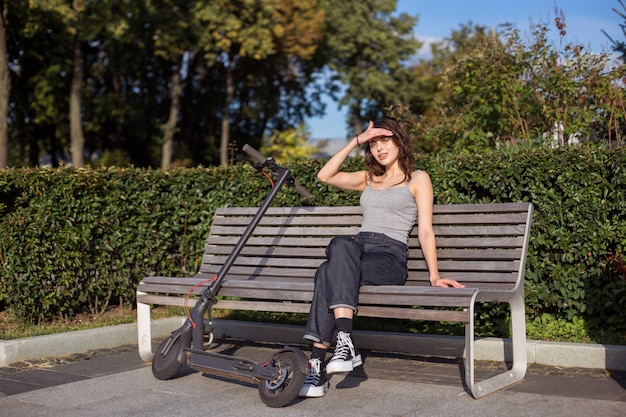 Foto elegante chica morena sentada en un banco con su scooter en un parque al aire libre en un clima soleado